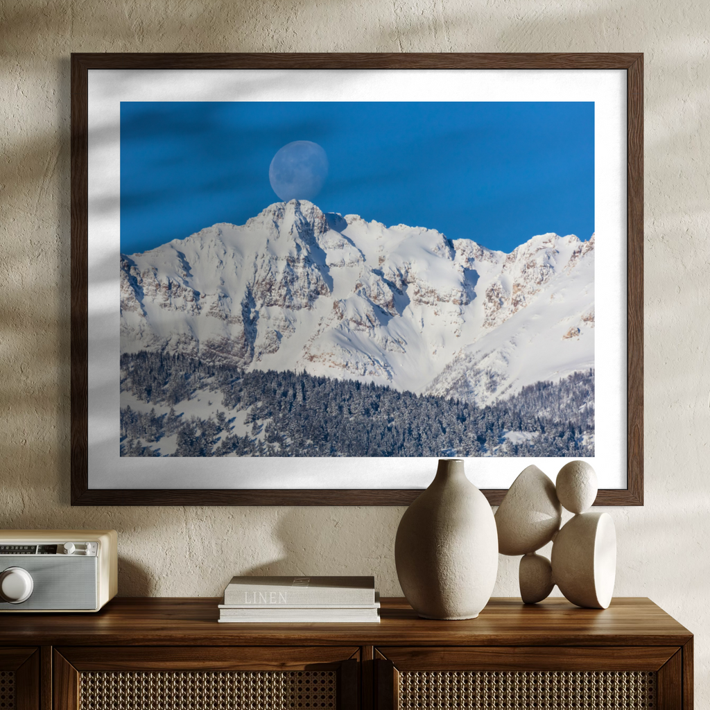 A framed wall art print showing a snowy mountain peak under a deep blue sky with a visible moon, displayed above a wooden console table with decorative vases and books in a styled interior setting.