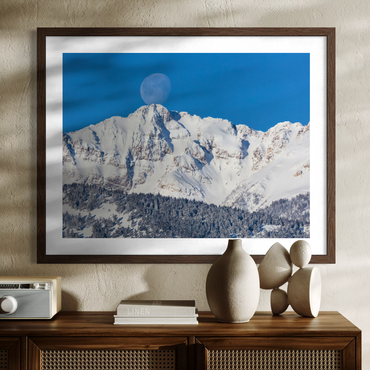 A framed wall art print showing a snowy mountain peak under a deep blue sky with a visible moon, displayed above a wooden console table with decorative vases and books in a styled interior setting.
