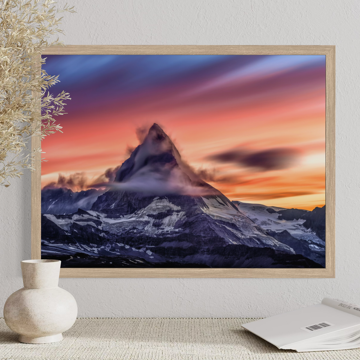 A framed wall art print showing a mountain peak at sunset, displayed on a textured wall above a table with a vase and an open book.