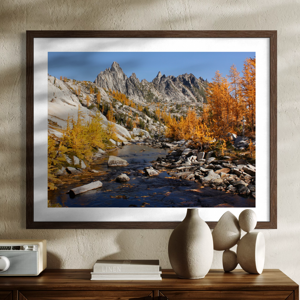 A wooden framed landscape art print depicting a rocky mountain stream with golden autumn trees, displayed on a wall above a wooden console table with decorative ceramic vases and books.