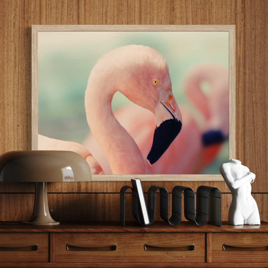 A wooden framed wall art print featuring a close-up flamingo portrait, displayed above a wooden console table with a lamp, books, and a small white figurine.