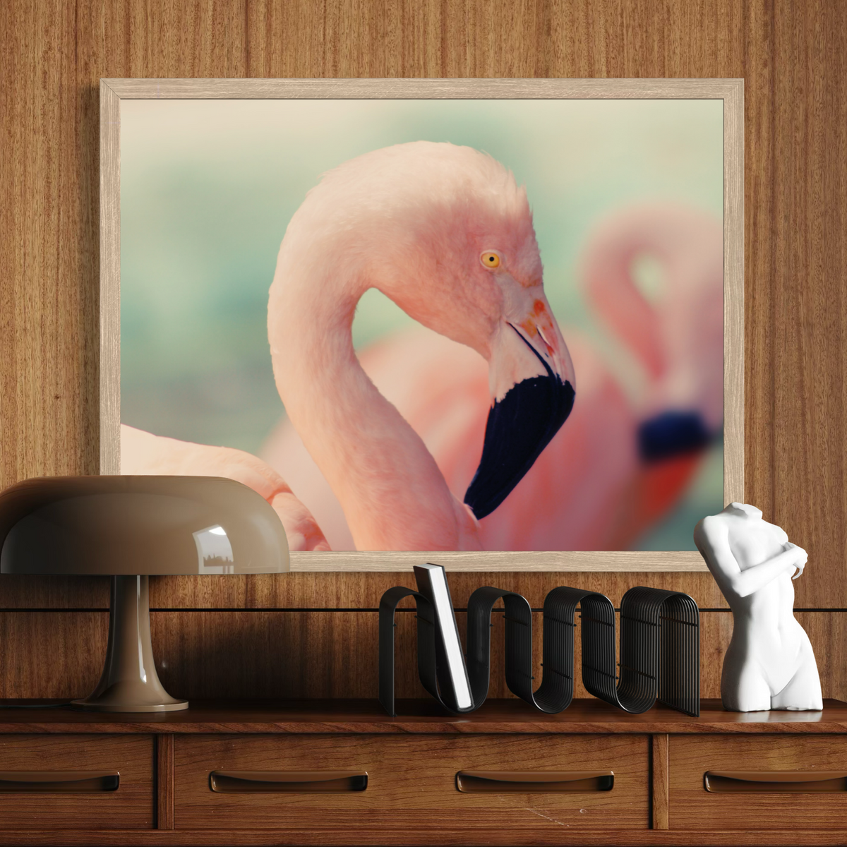 A wooden framed wall art print featuring a close-up flamingo portrait, displayed above a wooden console table with a lamp, books, and a small white figurine.