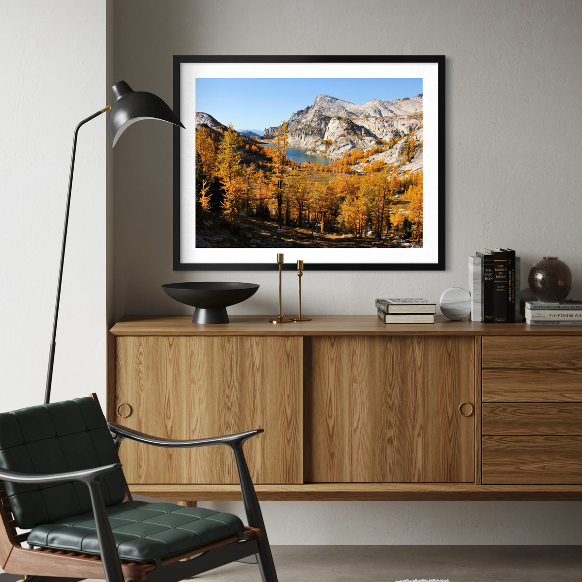 A black framed landscape art print of autumn mountains and golden trees, displayed on a gray wall above a wooden credenza in a modern living room setting.
