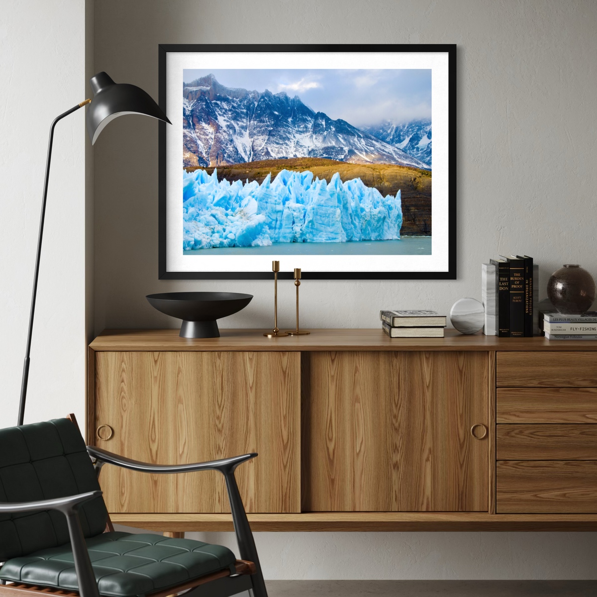 A framed landscape art print of a glacier and mountains displayed on a wall above a wooden credenza in a modern living room setting.