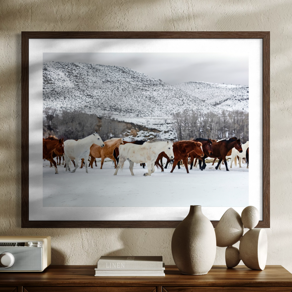 A framed wall art print showing a herd of horses walking through a snowy landscape, displayed on a wooden console table against a textured beige wall with decorative vases and a radio.