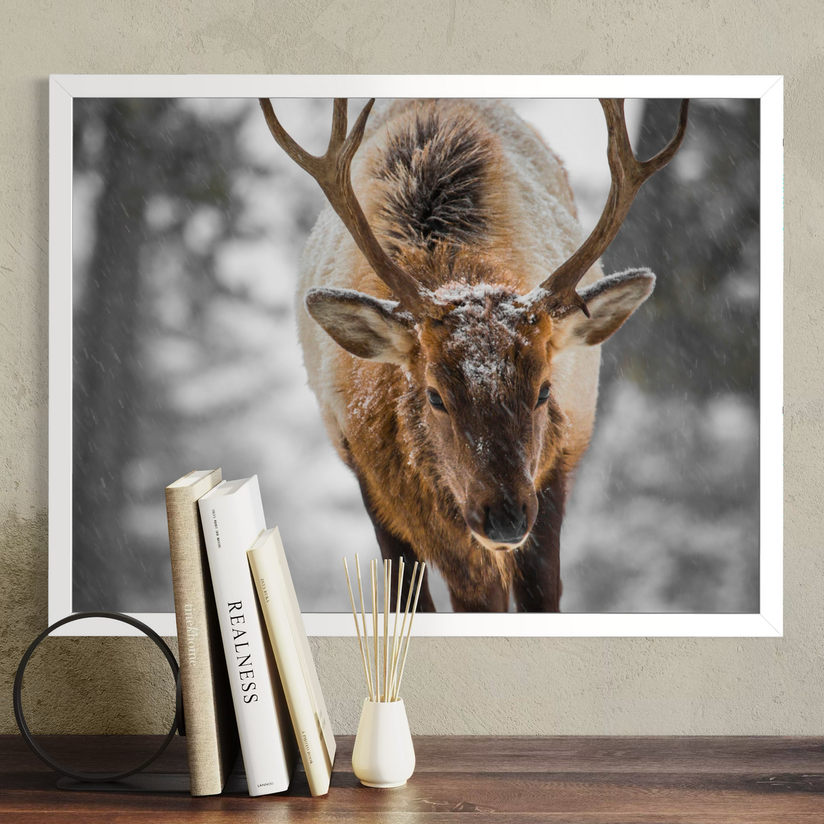 A white framed wall art print showing a close-up photograph of a deer with large antlers in a snowy outdoor scene, displayed above a wooden surface with books and a reed diffuser. The product being sold appears to be the framed print itself rather than the decor props.