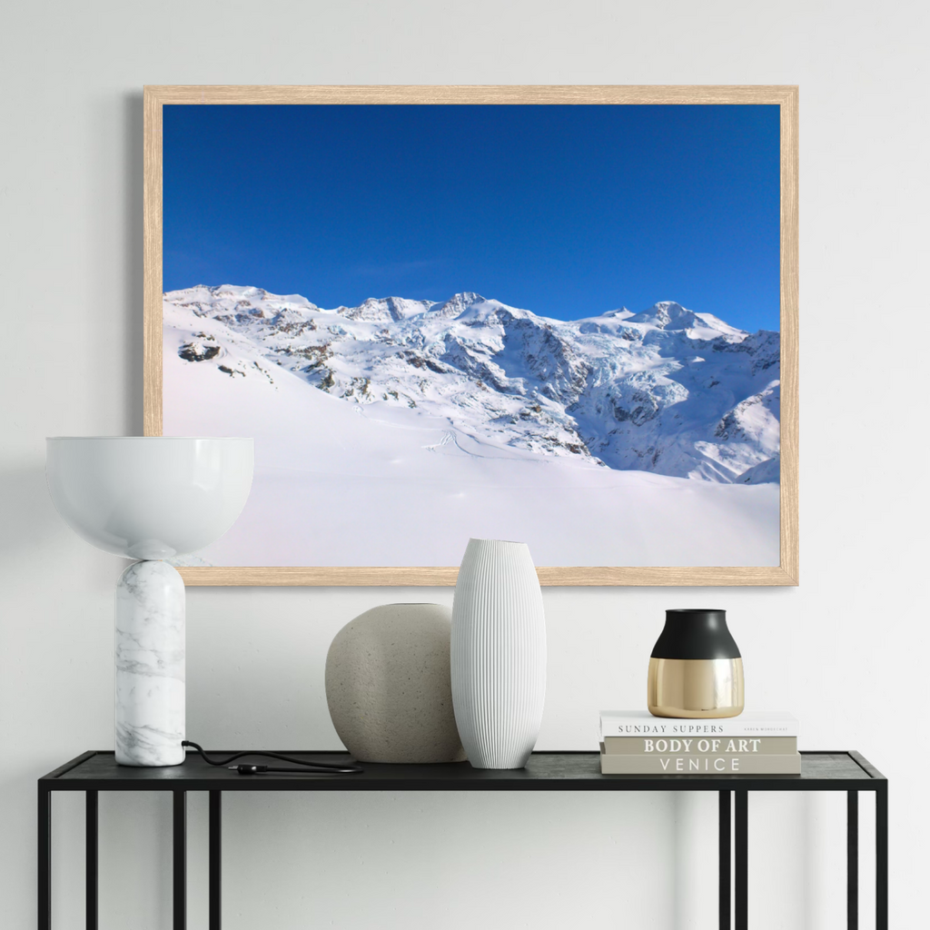A framed art print depicting a snowy mountain landscape under a clear blue sky, displayed on a light gray wall above a black console table with decorative vases and books.