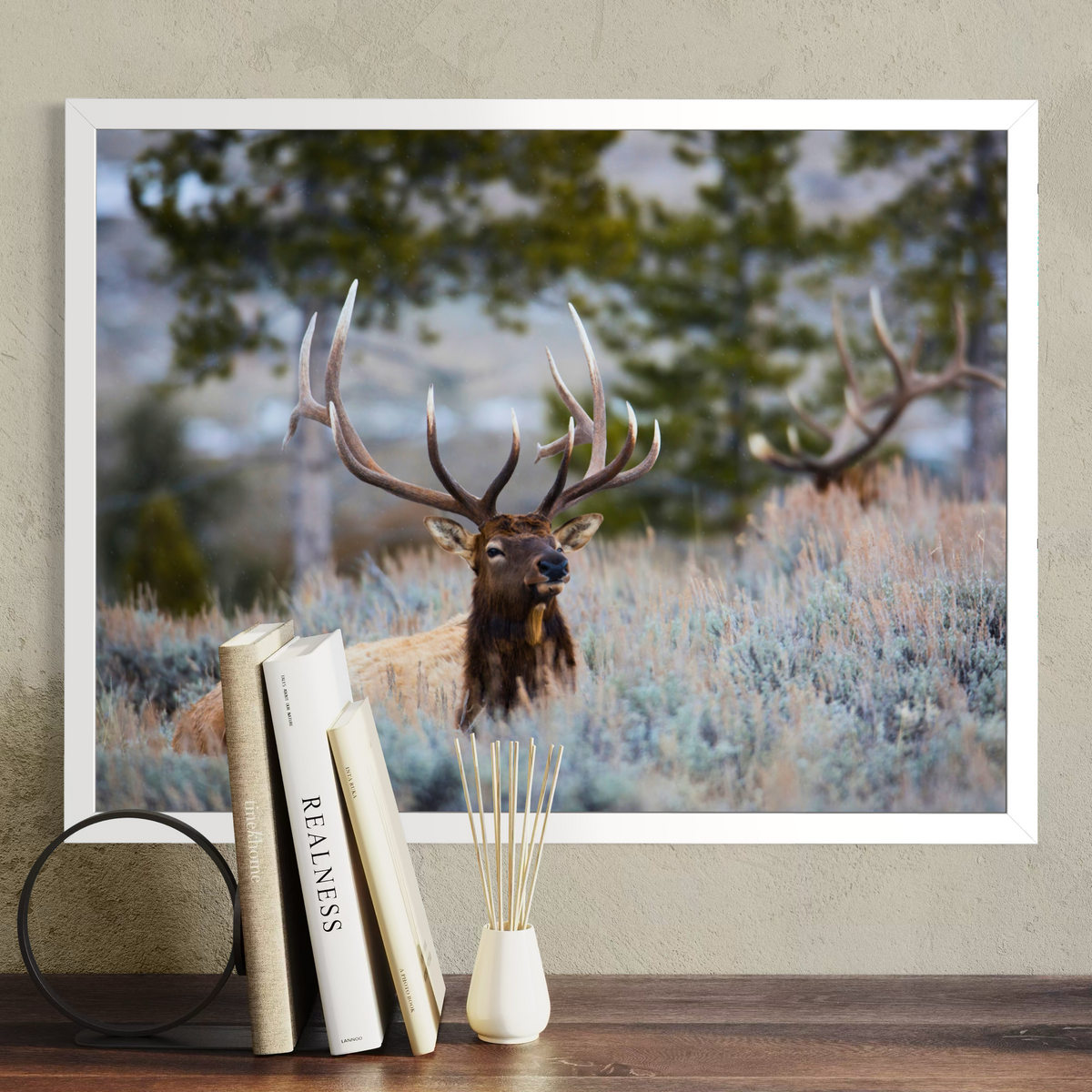 A framed wall art print showing a large elk deer with antlers in a grassy field, displayed above a wooden surface with books and a small vase in a styled interior setting.