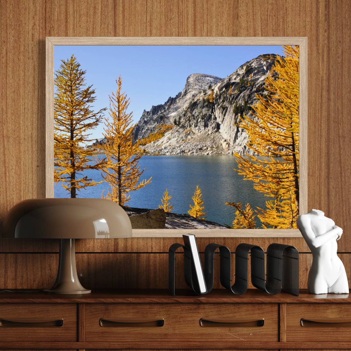 A wooden framed landscape art print depicting a mountain lake with golden trees, displayed on a wooden wall above a console table with a lamp, books, and a white sculpture.