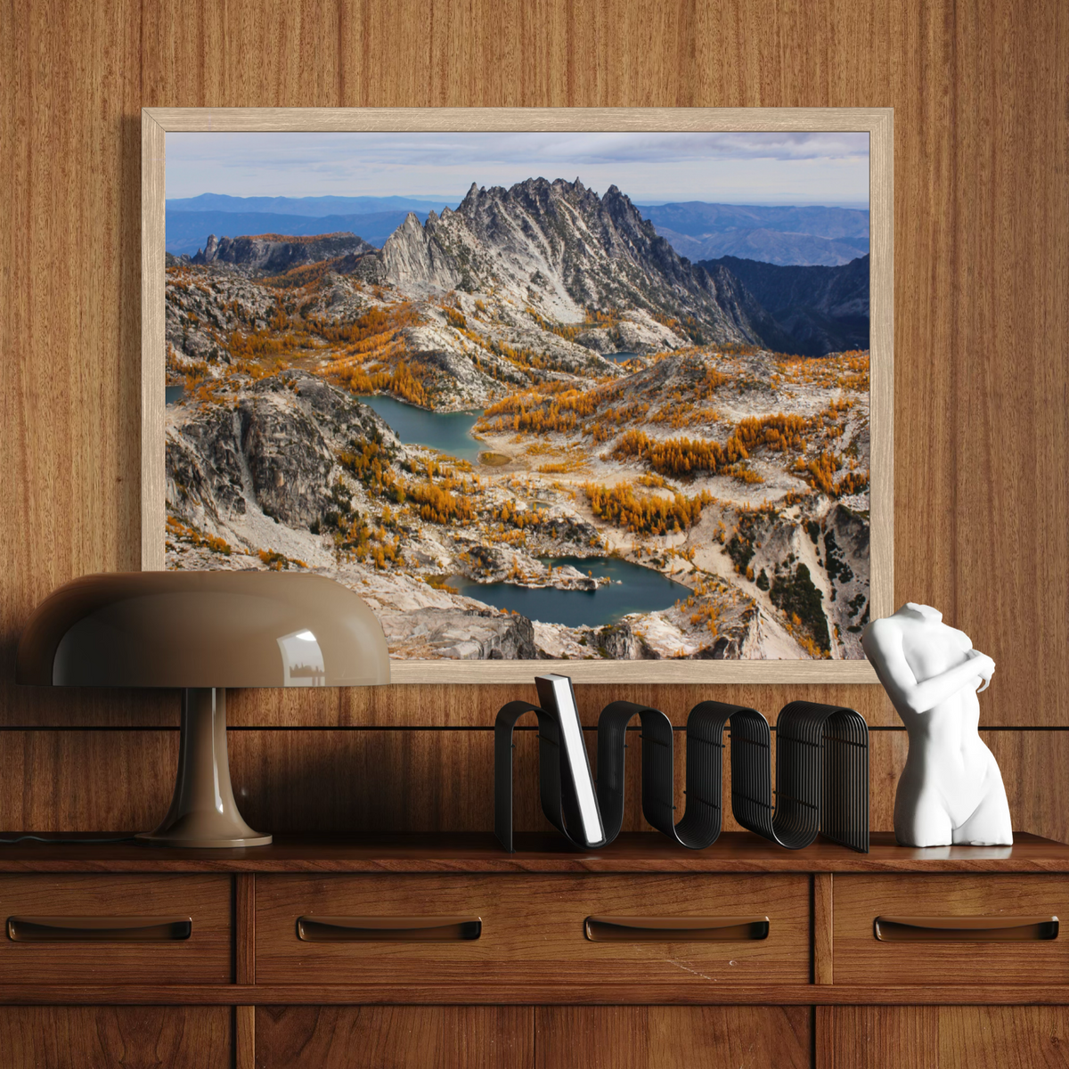 A wooden framed landscape art print depicting a rugged mountain range with blue lakes and golden autumn trees, displayed on a wooden console table above a lamp and decorative objects.