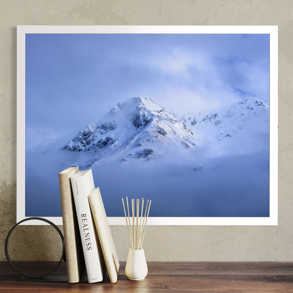 A framed wall art print showing a snowy mountain peak rising through blue clouds, displayed above a wooden surface with books, a black circular decor object, and a small white vase with reeds.