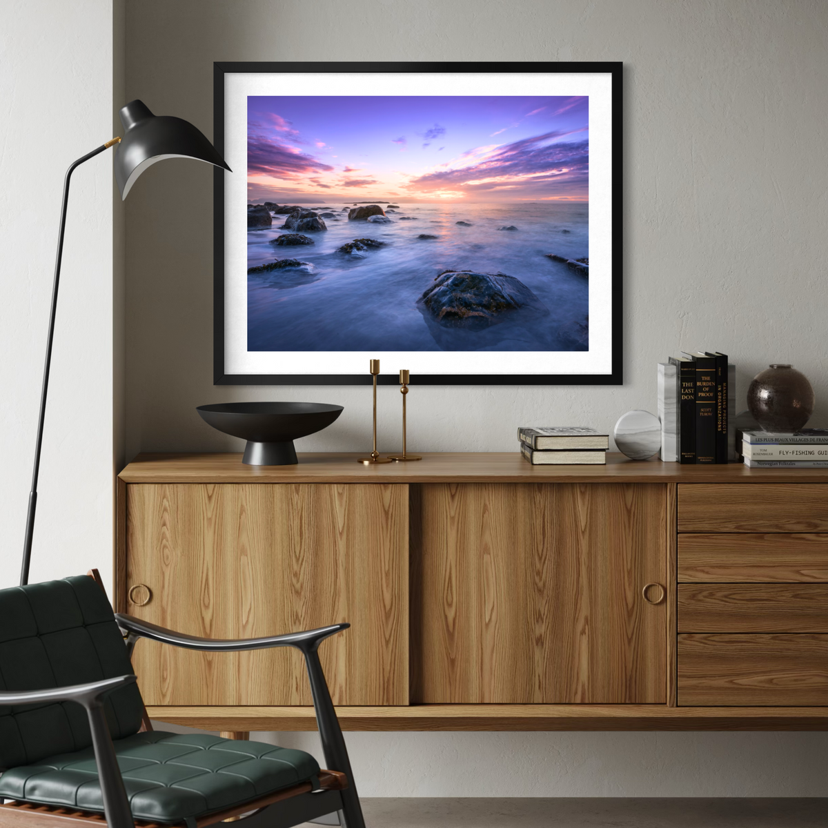 A framed wall art print of a purple and orange sunset over a rocky ocean, displayed on a light gray wall above a wooden credenza in a modern living room setting.