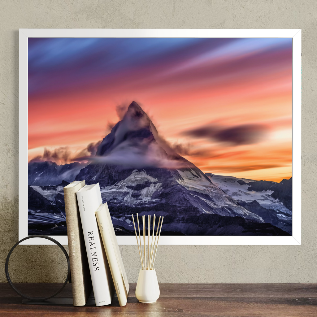 A framed wall art print showing a dramatic mountain peak at sunset with pink, orange, purple, and blue sky, displayed above a wooden surface with books and a small vase in a styled interior setting.