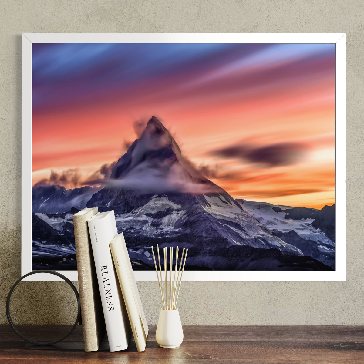 A framed wall art print showing a dramatic mountain peak at sunset with pink, orange, purple, and blue sky, displayed above a wooden surface with books and a small vase in a styled interior setting.