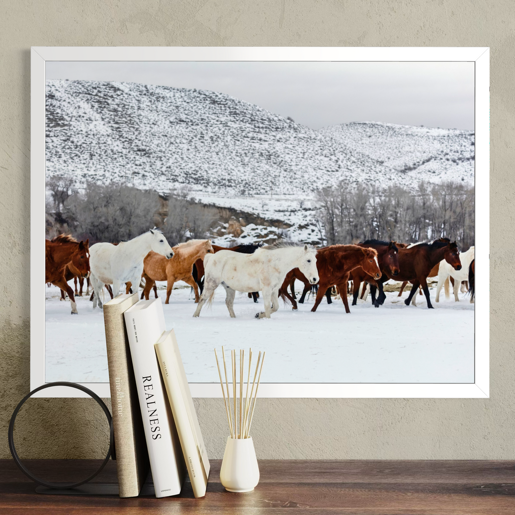 A framed wall art print showing a herd of wild horses walking through a snowy landscape with mountains in the background, displayed on a wall above a wooden surface with books and a diffuser.