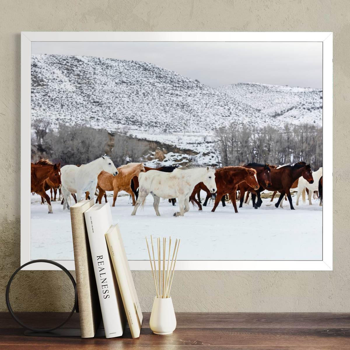 A framed wall art print showing a herd of wild horses walking through a snowy landscape with mountains in the background, displayed on a wall above a wooden surface with books and a diffuser.