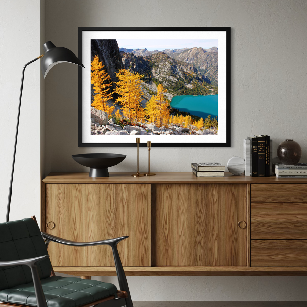 A black wooden framed landscape art print featuring golden autumn larch trees and a turquoise lake in mountains, displayed on a light gray wall above a wooden credenza in a modern living room setting.