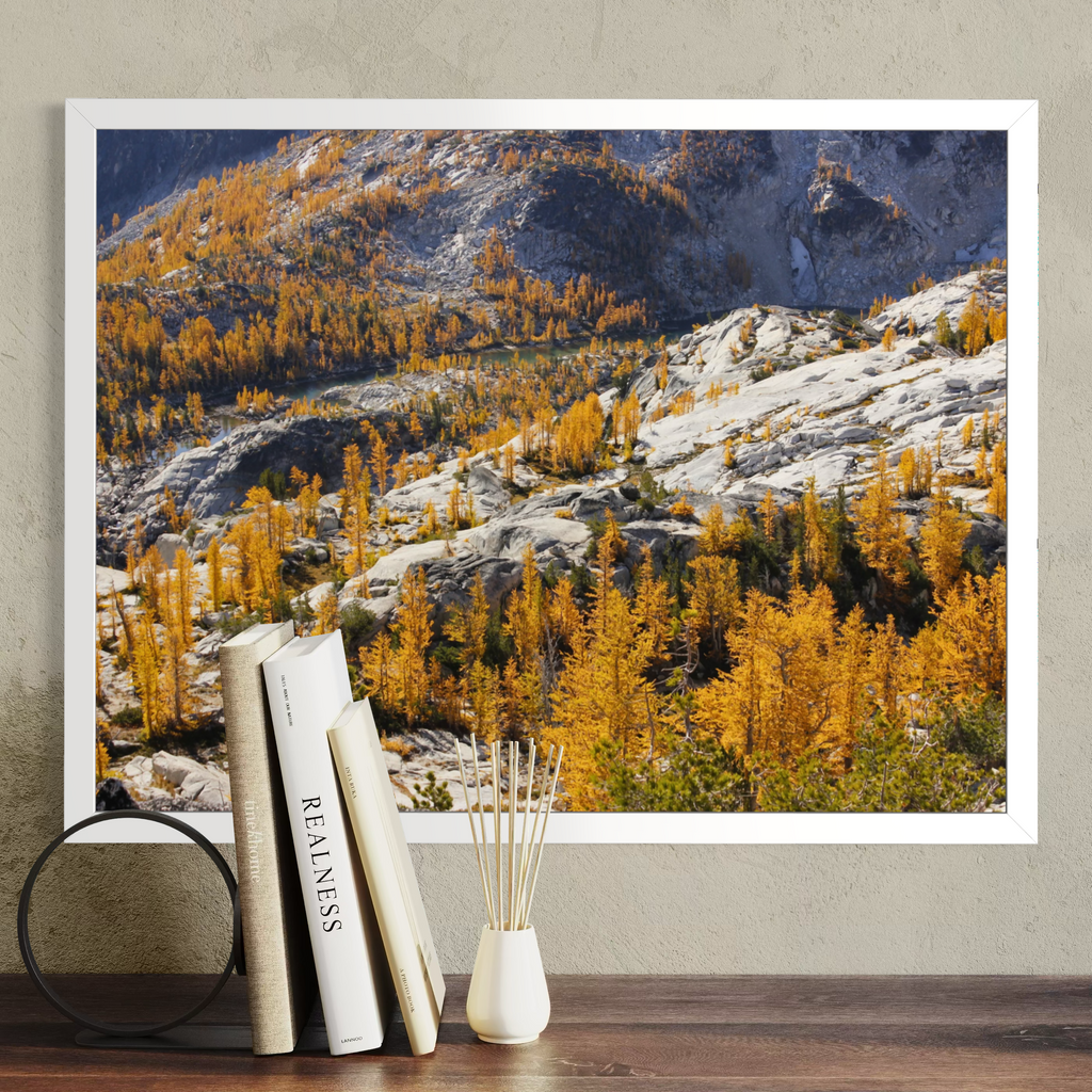A white framed wall art print showing a mountain landscape with golden yellow larchwood trees and rocky slopes, displayed above a wooden surface with books and a reed diffuser in a styled interior setting.