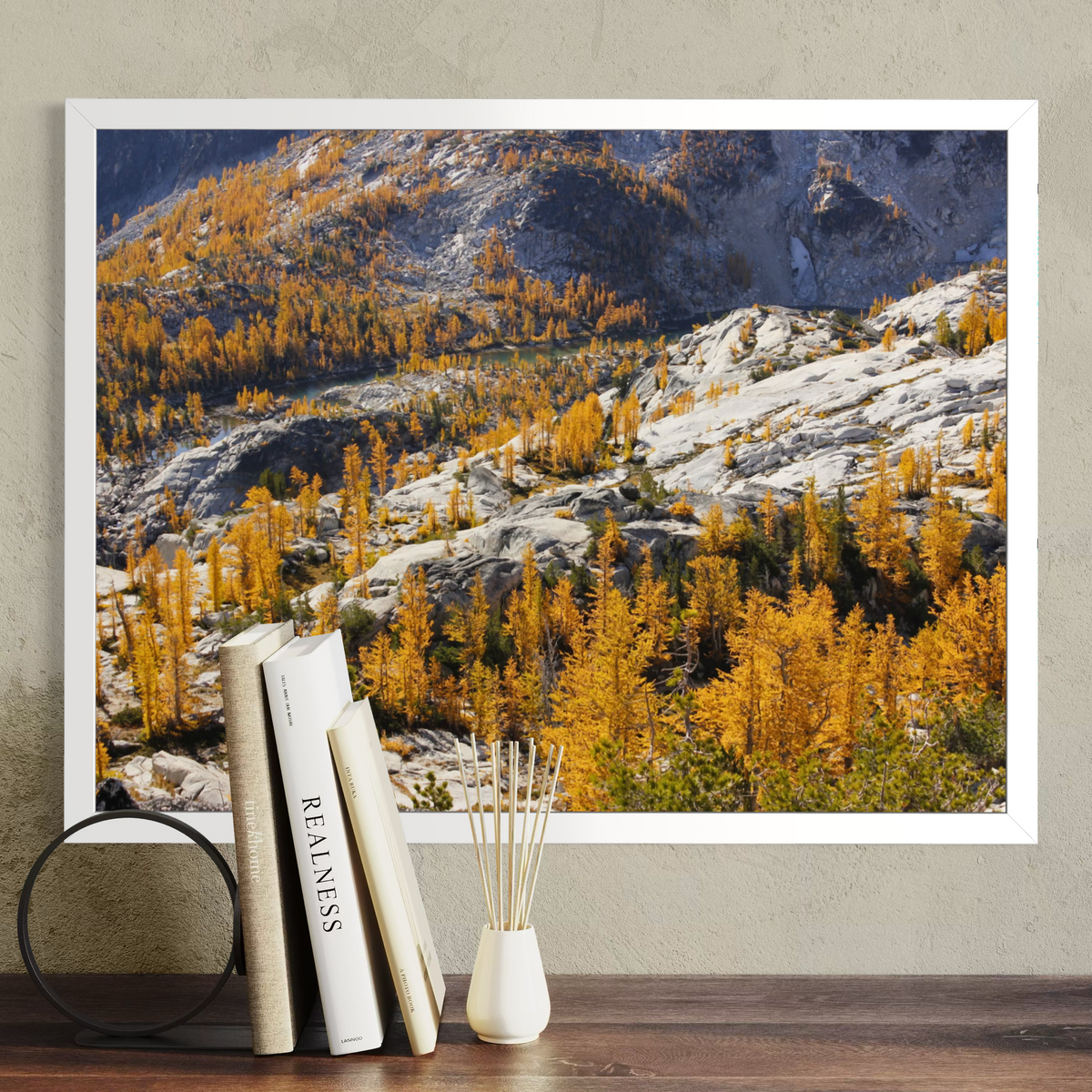 A white framed wall art print showing a mountain landscape with golden yellow larchwood trees and rocky slopes, displayed above a wooden surface with books and a reed diffuser in a styled interior setting.