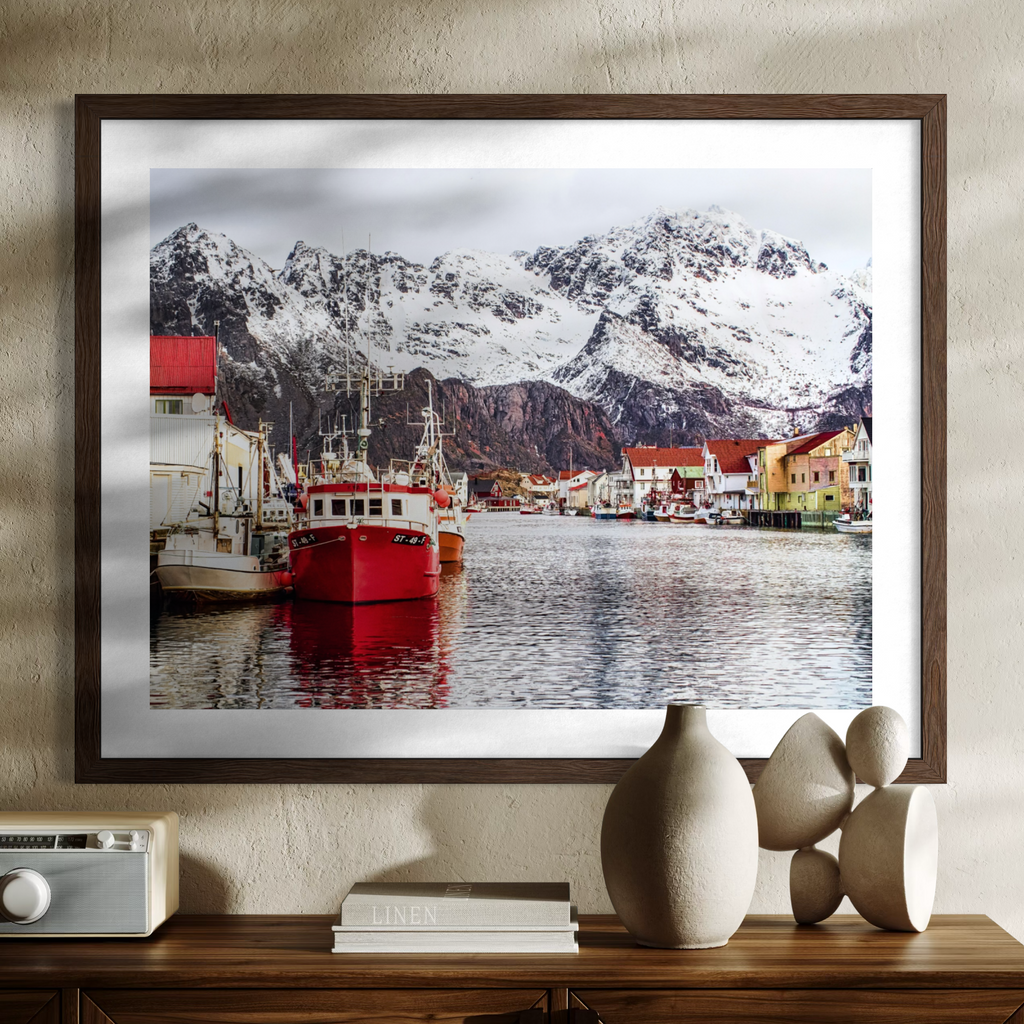 A framed art print depicting a scenic harbor with snow-covered mountains, colorful boats, and a small village, displayed on a wooden console table with decorative vases and books against a textured beige wall.