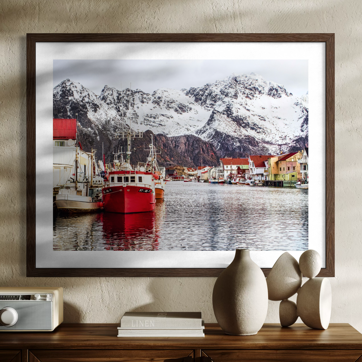 A framed art print depicting a scenic harbor with snow-covered mountains, colorful boats, and a small village, displayed on a wooden console table with decorative vases and books against a textured beige wall.