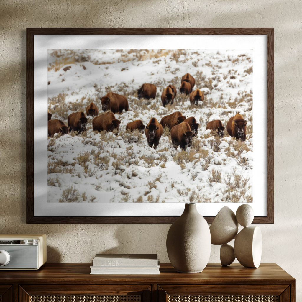 A wooden framed wall art print showing a herd of brown bison walking through a snowy field, displayed above a wooden console table with decorative vases and books against a textured beige wall.