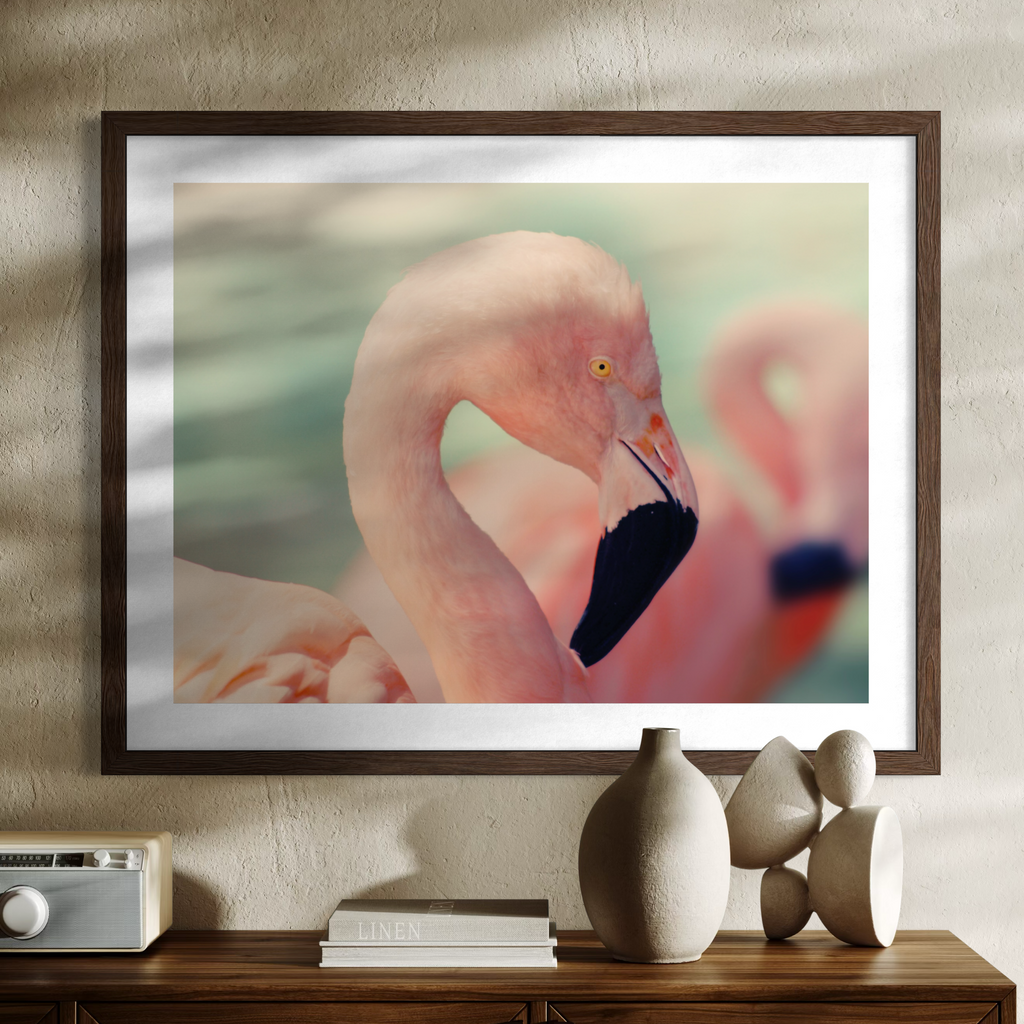 A wooden framed art print featuring a close-up photograph of a pink flamingo, displayed on a textured beige wall above a wooden console table with decorative ceramic vases and books.