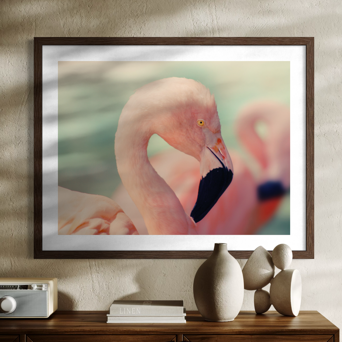 A wooden framed art print featuring a close-up photograph of a pink flamingo, displayed on a textured beige wall above a wooden console table with decorative ceramic vases and books.