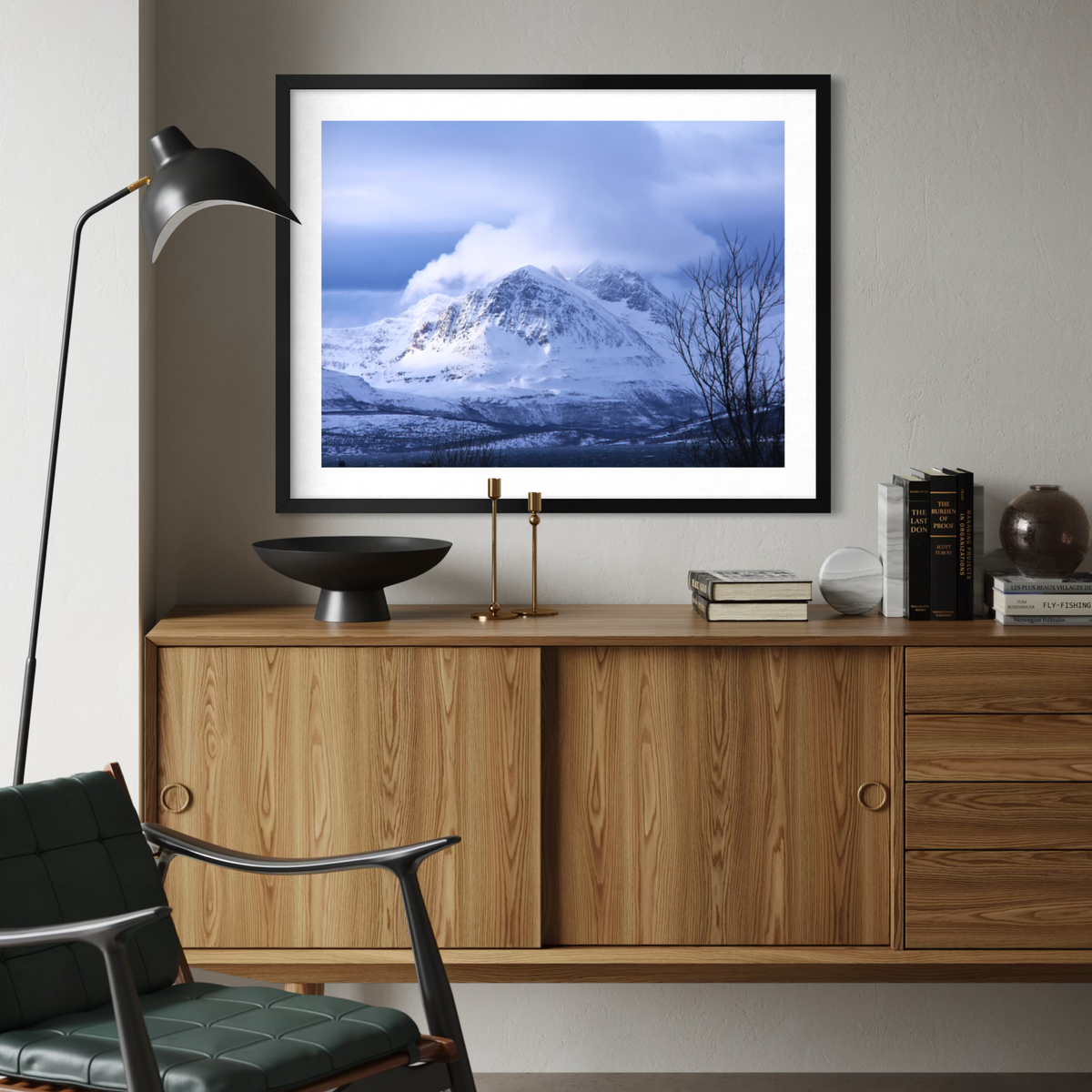 A framed landscape art print of a snowy mountain under a cloudy blue sky, displayed on a light gray wall above a wooden credenza in a modern living room setting.