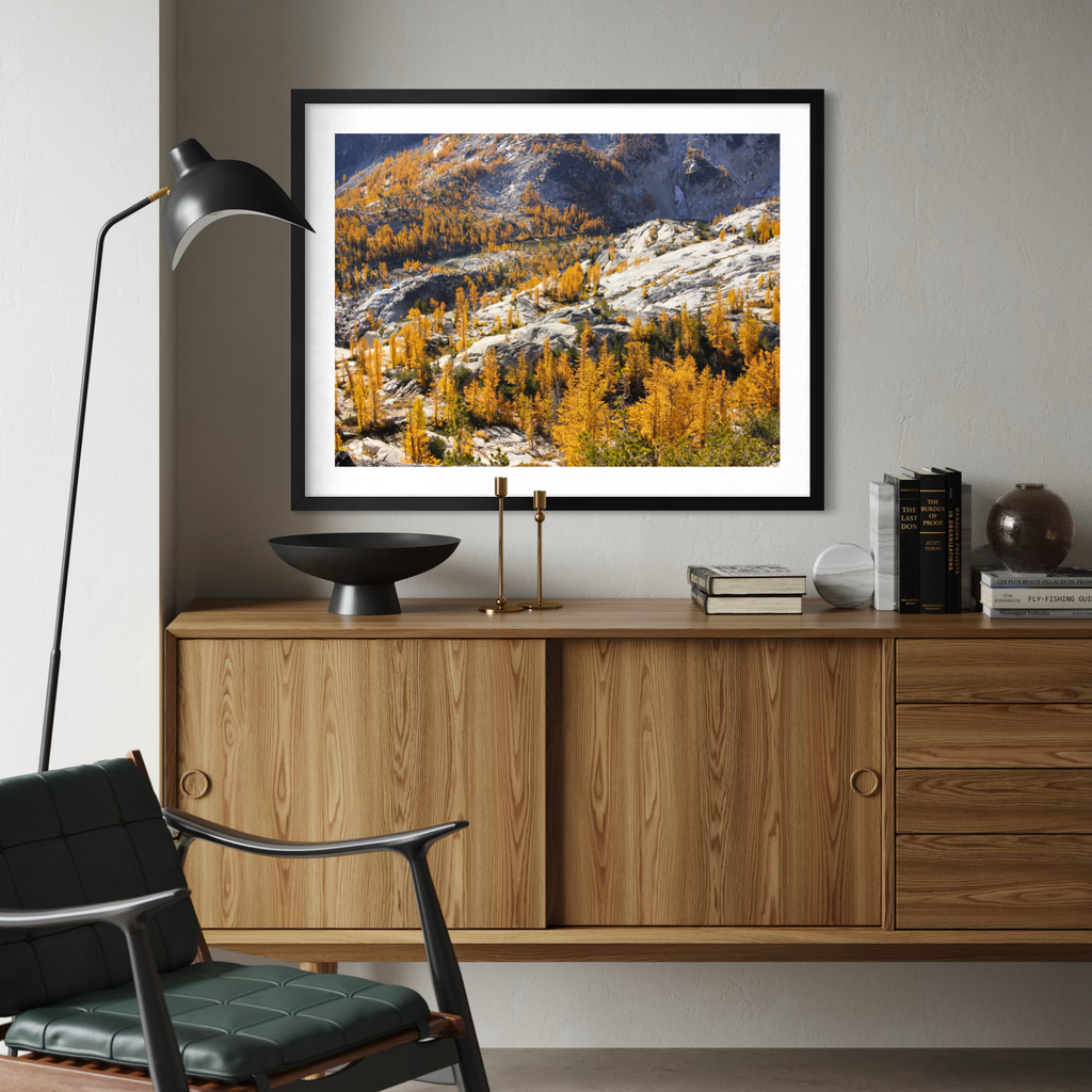 A black framed art print depicting a mountain landscape with golden yellow larchwood trees and blue-gray mountains, styled on a light gray wall above a wooden credenza in a modern living room setting.