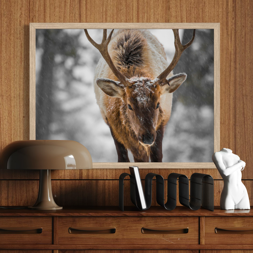 A framed wall art print showing a close-up photograph of a deer elk with antlers in a snowy outdoor scene, displayed above a wooden console table with a lamp, books, and a small white figurine.