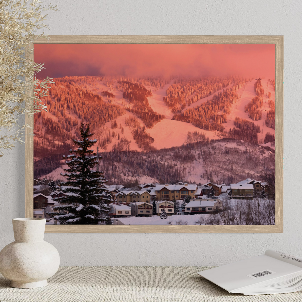 A framed wall art print showing a snowy mountain village at sunset, displayed on a tabletop against a light wall with a vase and book nearby.