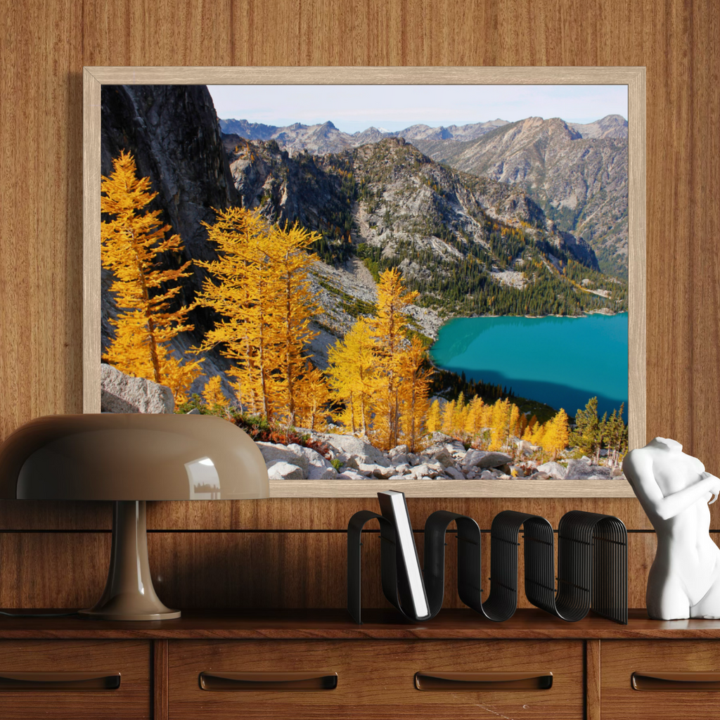 A wooden framed landscape art print depicting golden larch trees, rocky mountains, and a turquoise lake, displayed on a wooden wall above a desk with a lamp, books, and a white bust.