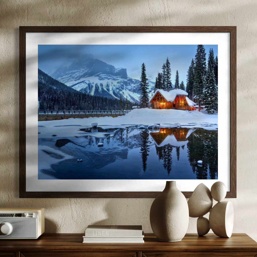 A framed wall art print showing a snowy mountain landscape with a glowing cabin reflected in a calm lake, displayed above a wooden console with decorative objects.
