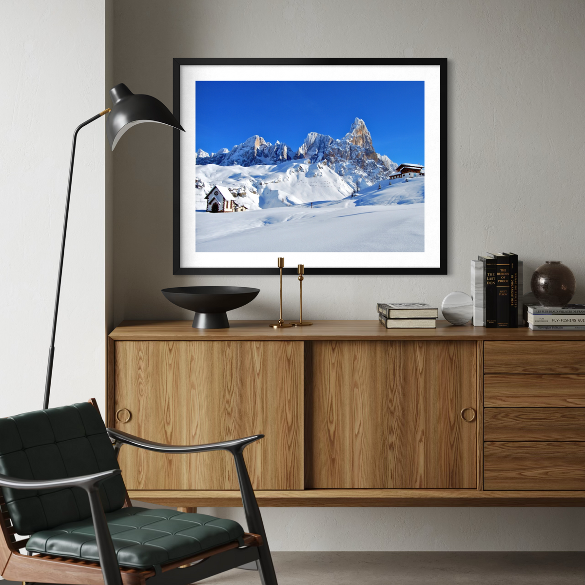 A framed art print depicting a snowy mountain landscape under a clear blue sky, displayed on a light gray wall above a mid-century modern wooden credenza in a styled living room setting.