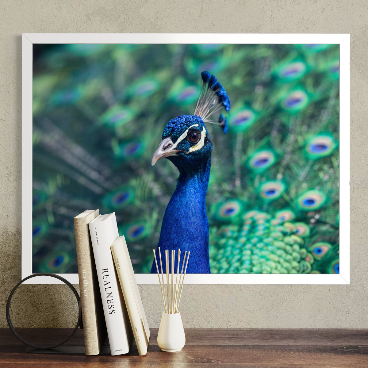 A framed wall art print featuring a close-up photograph of a peacock with vivid blue and green feathers, displayed above a wooden surface with books and a reed diffuser. The frame is mounted on a textured beige wall.