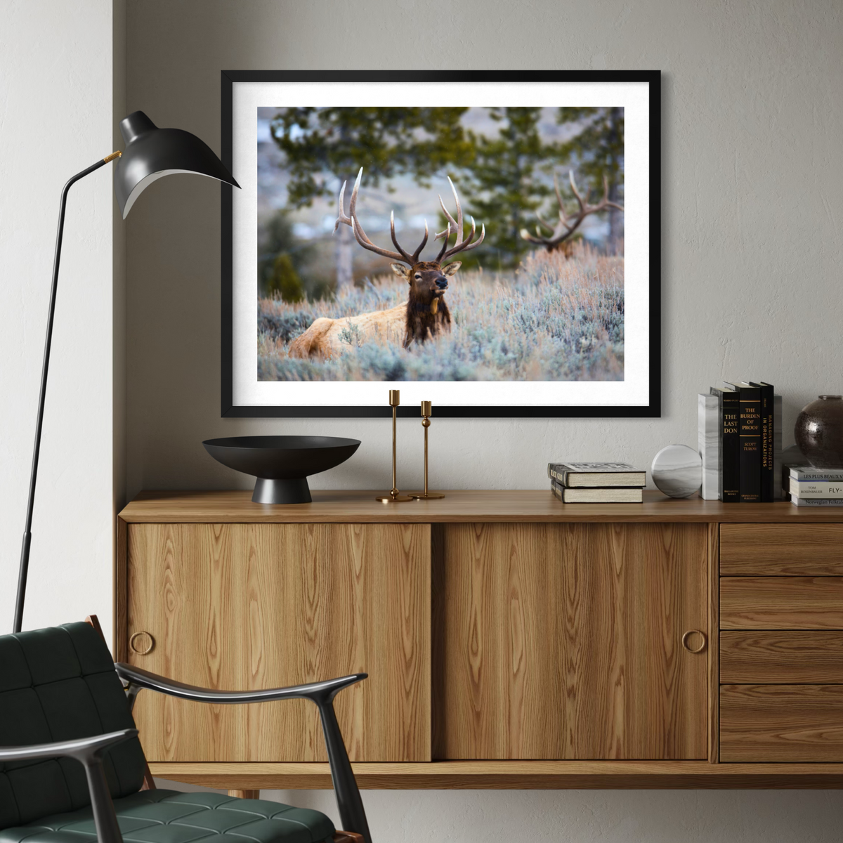 A framed art print depicting a majestic stag with large antlers resting in a field, displayed on a light gray wall above a wooden sideboard with a black floor lamp and a green armchair in the foreground.