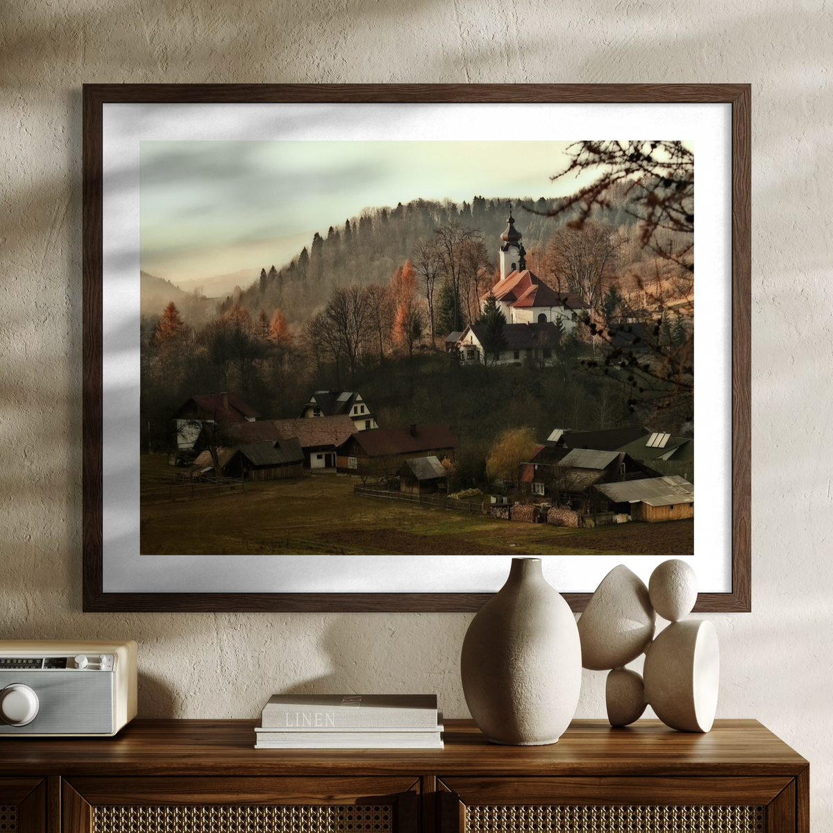 A framed art print depicting a scenic mountain village with a church and autumn trees, styled on a wooden console table with decorative ceramic vases and books against a textured beige wall.