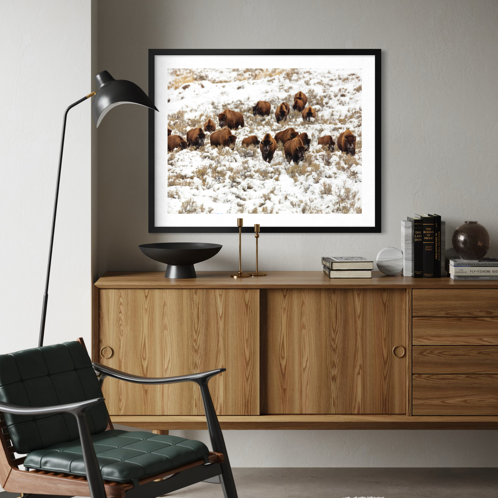 A black framed art print depicting a herd of bison in a snowy landscape, displayed on a light gray wall above a mid-century modern wooden credenza in a styled living room setting.
