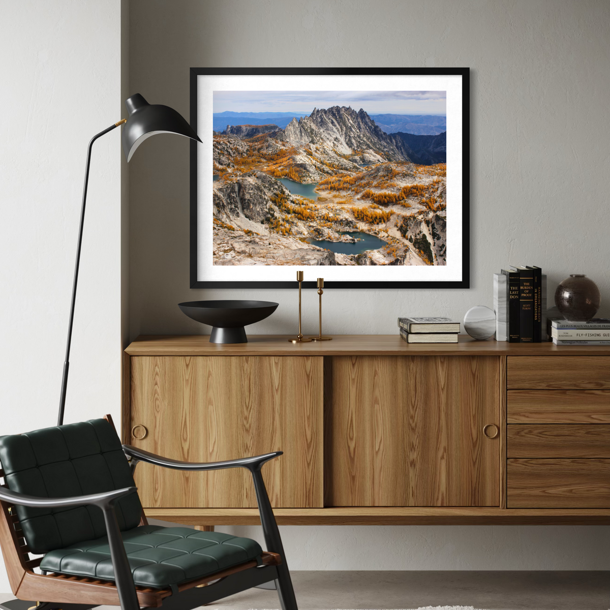 A black framed landscape art print of a mountain range with autumn foliage and a river, displayed on a gray wall above a wooden credenza in a modern living room setting.
