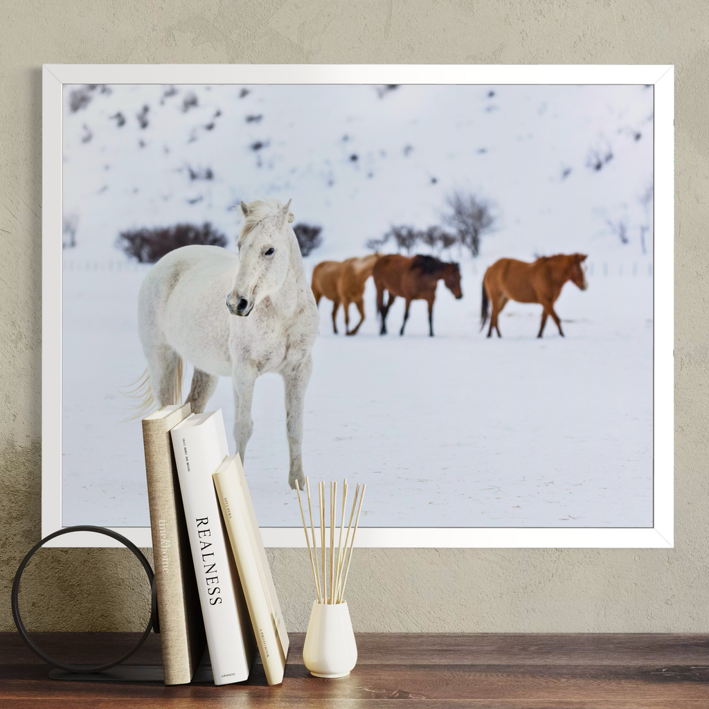 A framed wall art print showing a snowy landscape with a white horse in the foreground and two brown horses behind it, displayed above a wooden surface with books and a small vase.