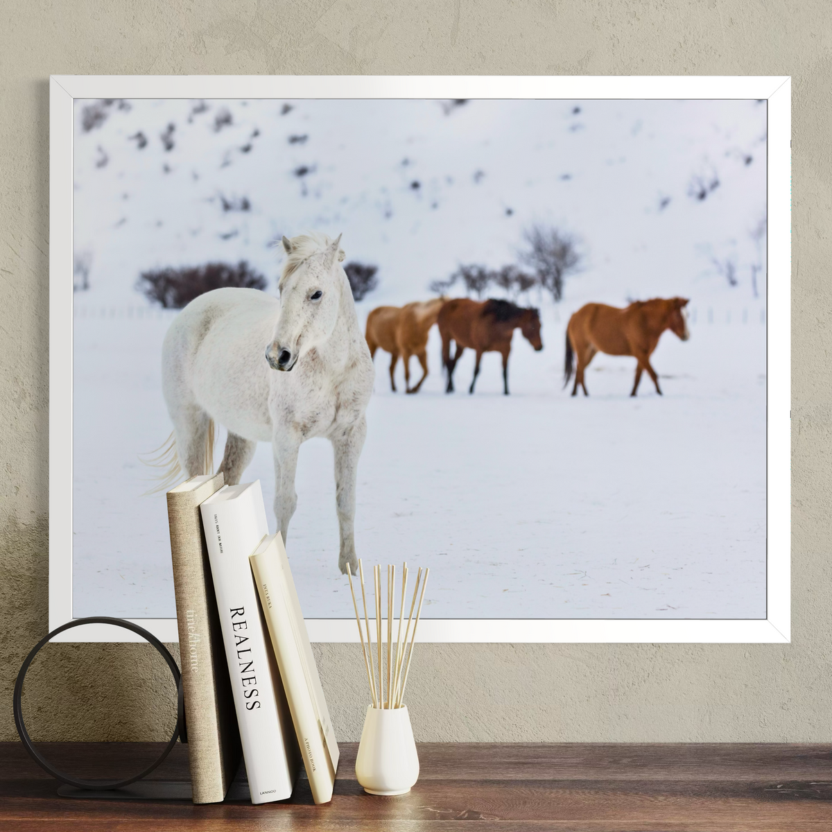 A framed wall art print showing a snowy landscape with a white horse in the foreground and two brown horses behind it, displayed above a wooden surface with books and a small vase.