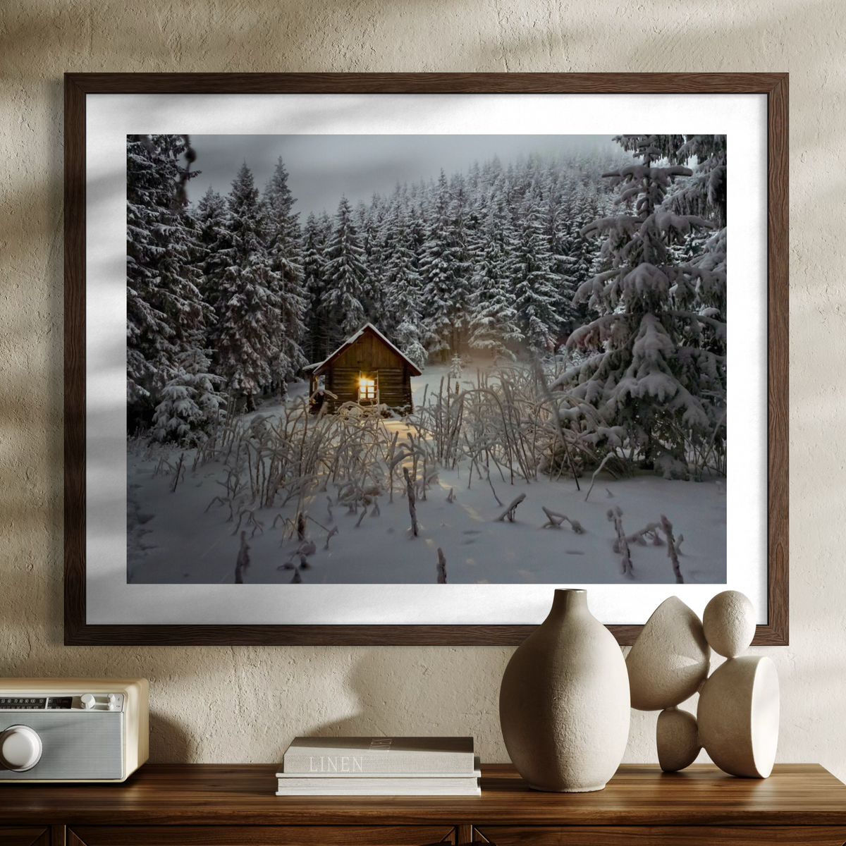 A framed winter landscape photograph hanging on a beige wall above a wooden console table. The print shows a snow-covered forest with a small cabin lit from within.