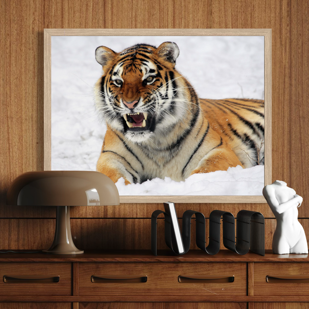 A wooden framed wall art print depicting a roaring tiger in white snow, mounted on a wood-paneled wall above a wooden console table with a lamp, books, and a small white figurine.