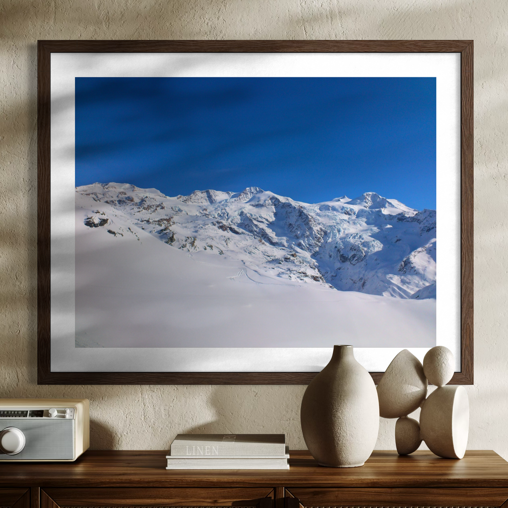 A framed art print depicting a snowy mountain landscape under a deep blue sky, displayed on a textured beige wall above a wooden console table with decorative vases and a radio.