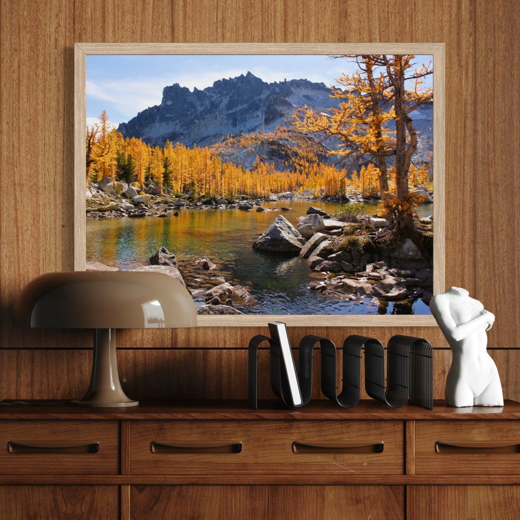 A wooden framed landscape art print depicting a mountain lake with golden autumn trees, displayed on a wooden wall above a cabinet with a lamp, books, and a white sculpture.