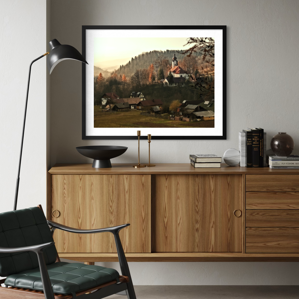 A framed landscape art print depicting a quaint village with a church on a hill, styled on a gray wall above a wooden sideboard in a modern living room setting.