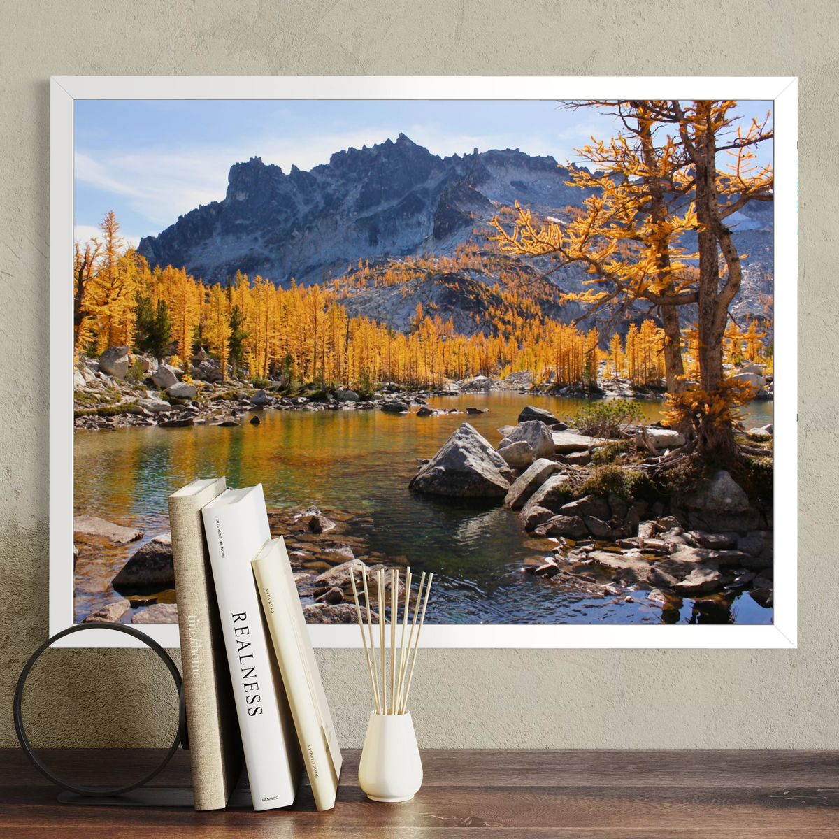 A white framed landscape art print depicting a mountain, golden larchwood trees, and a rocky lake, styled on a shelf next to books and a small vase.