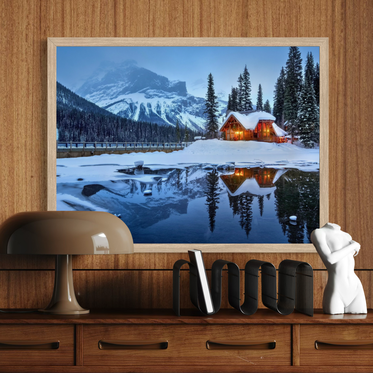 A framed landscape art print depicting a snowy mountain cabin at dusk, hung on a wooden wall above a console table with a lamp, books, and a small white sculpture.