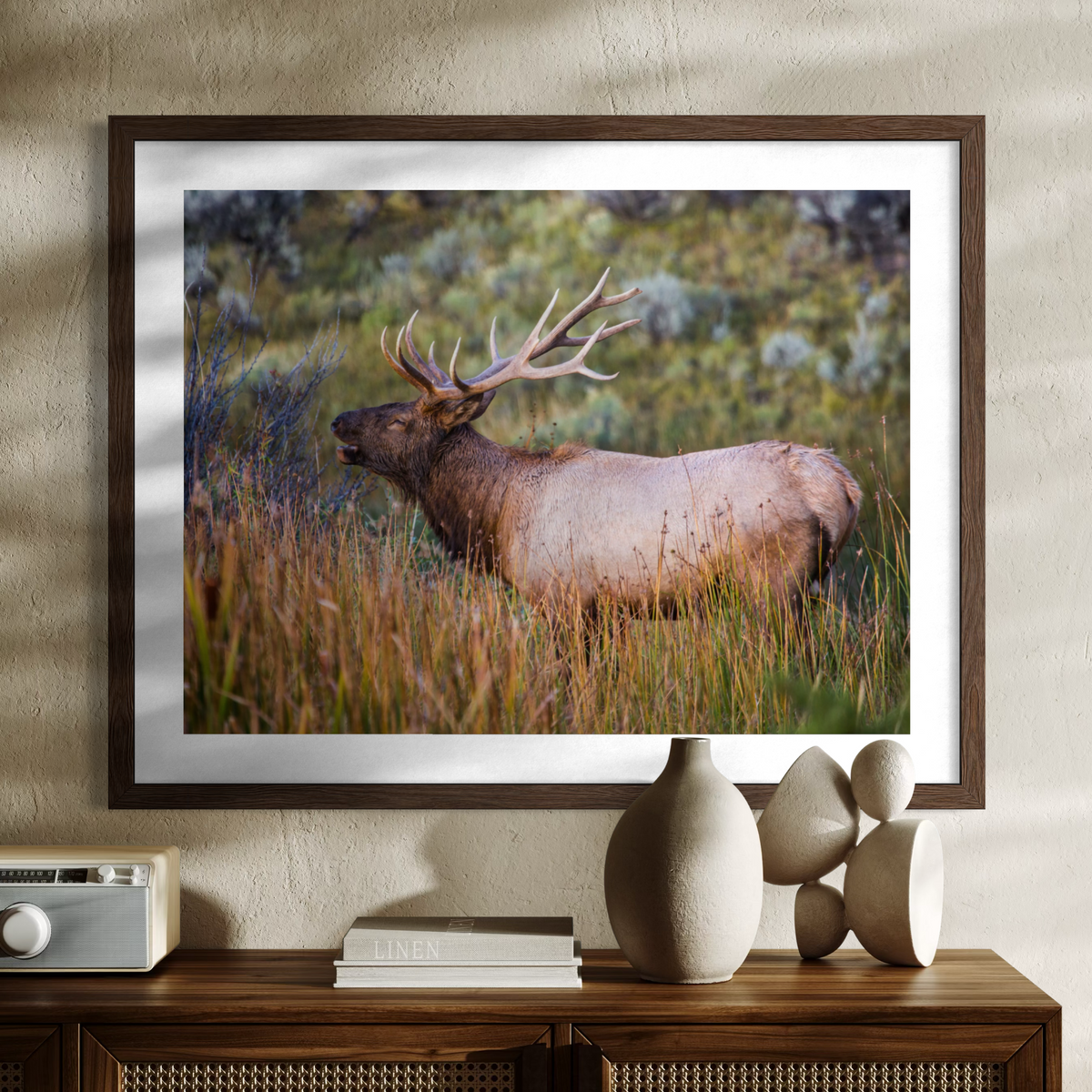 A wooden framed wall art print showing a large elk standing in tall grass, displayed above a wooden console table with decorative vases and books against a textured beige wall.